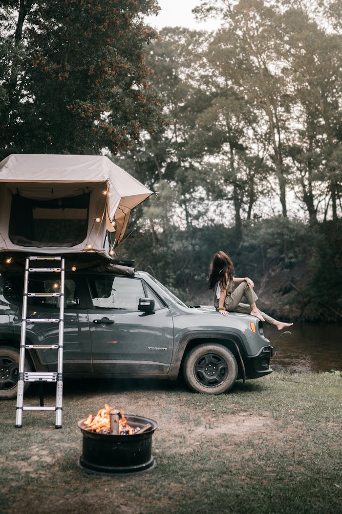Home A woman sits on a jeep by a campfire, enjoying an outdoor adventure.